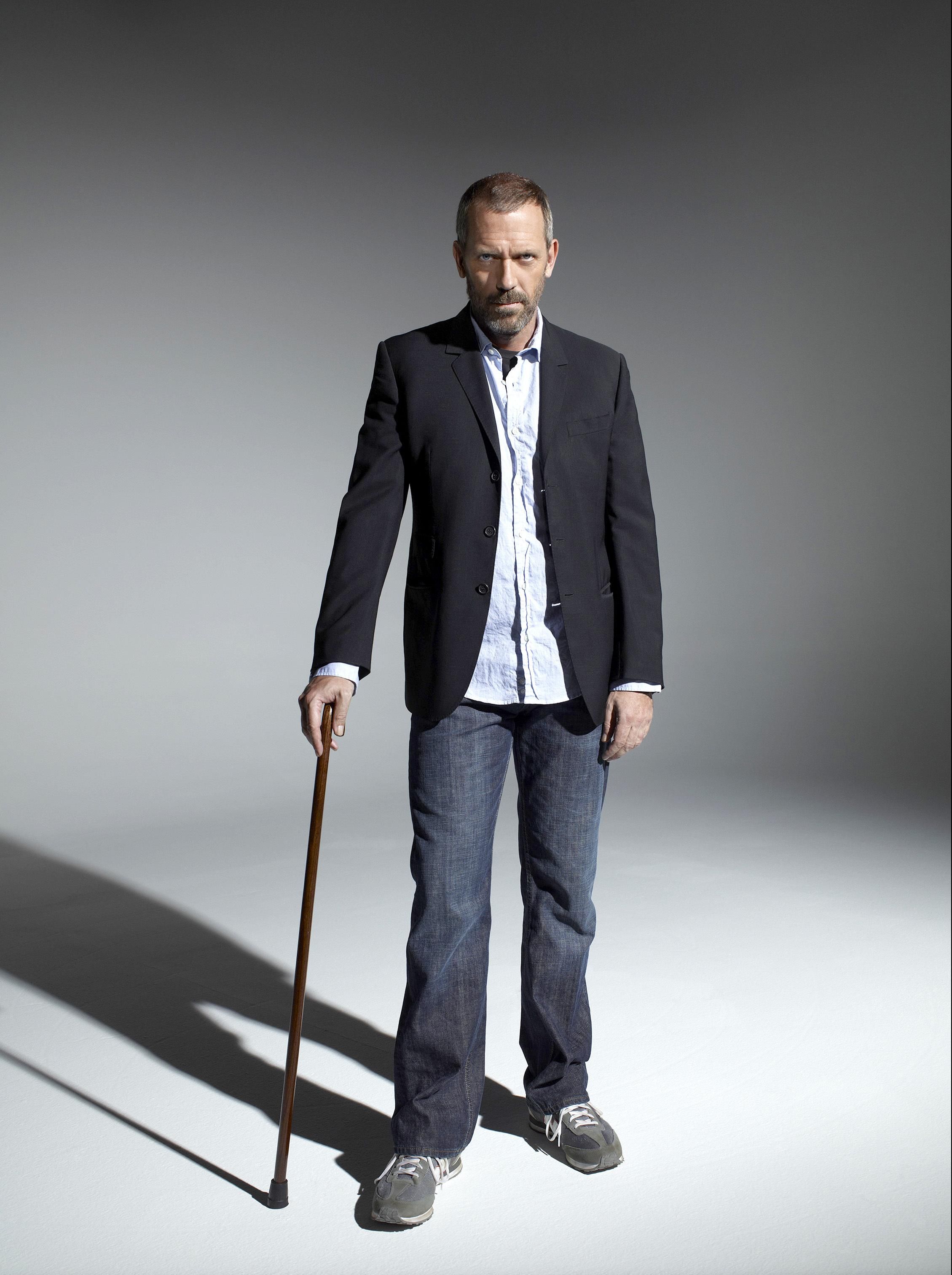 Actor named Hugh Laurie is standing with the walking stick. The background is grey, the actor wears black jacket, light blue shirt, dark jeans and sneakers. The photo is from photoshoot to Dr House.