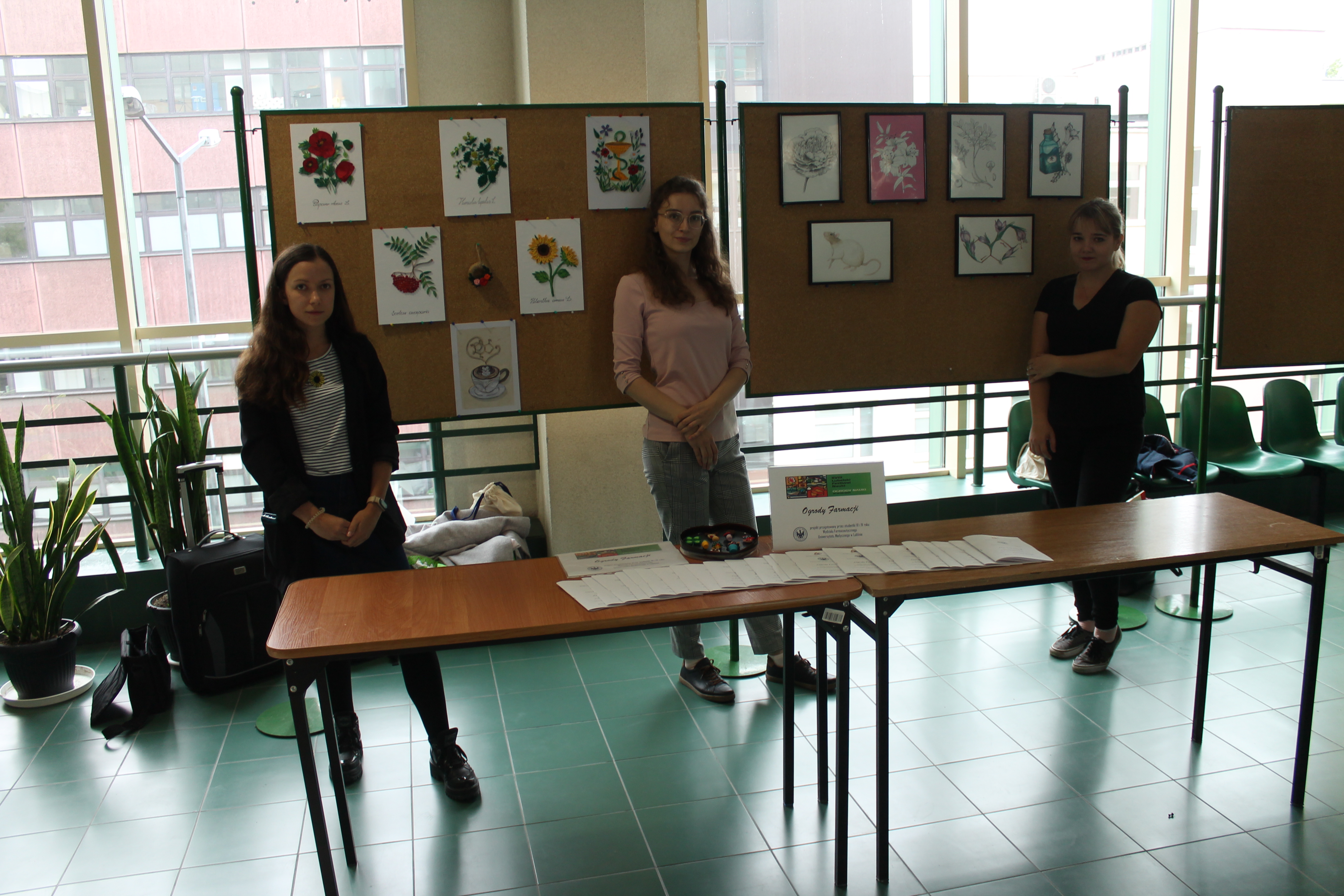 cztery dziewczyny stoją na tle tablicy z pracami plastycznymi, a za stołem, na którym leżą foldery promocyjne
