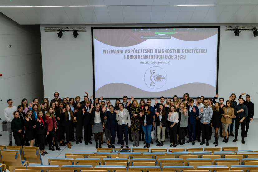 Organizatorzy i uczestnicy konferencji stoją w grupie na dole auli i machają do obiektywu.