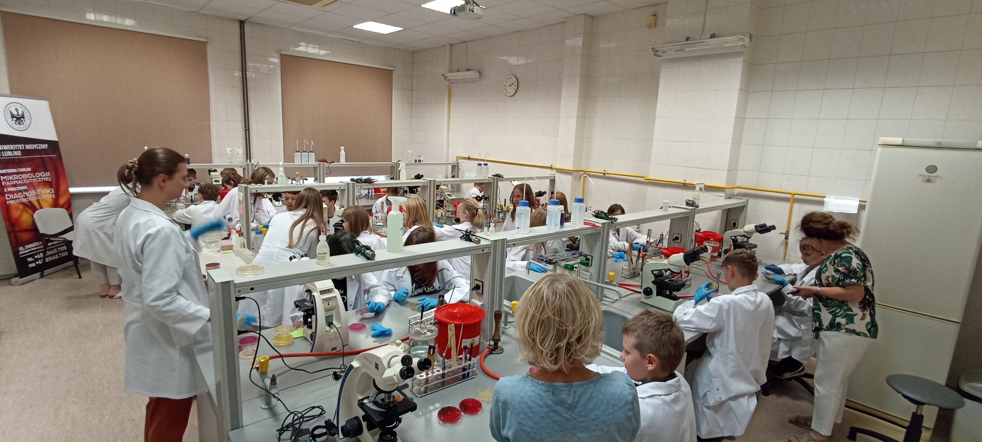W laboratorium mikrobiologicznym jest dużo osób przy stanowiskach - studenci, dzieci i dydaktycy.